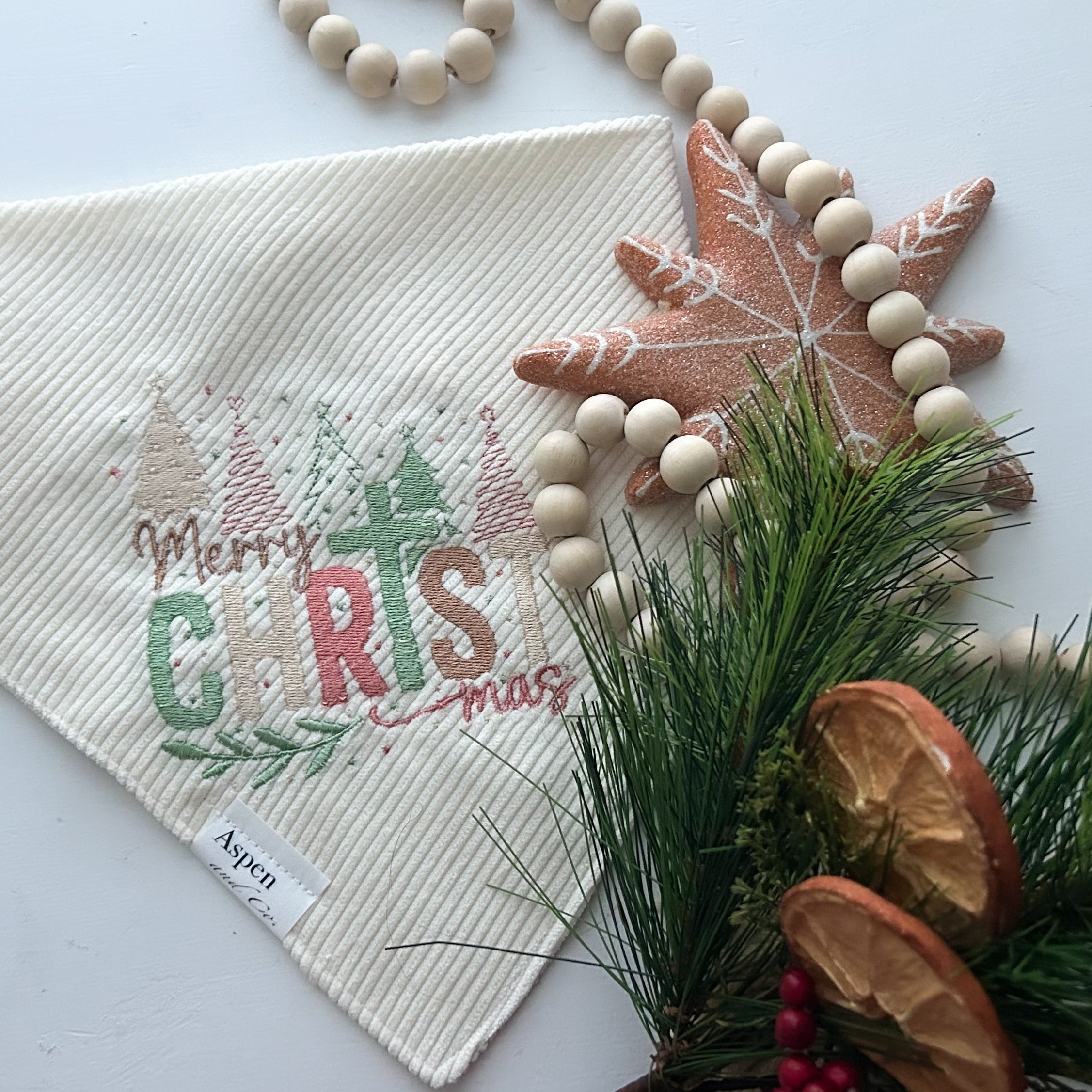 Cream corduroy dog bandana embroidered with “Merry ChrisTmas” in pastel green, red, and gold thread, featuring Christmas trees and festive details. Snap-on handmade bandana by Aspen & Co., styled with holiday props.