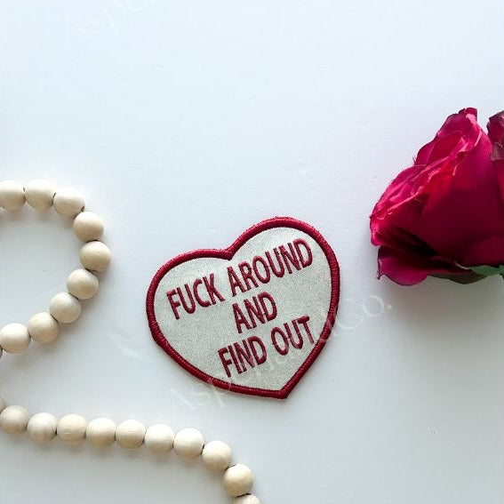 Glitter Heart-shaped patch with text 'F**K AROUND AND FIND OUT' on a white background with a pink rose and wooden beads.