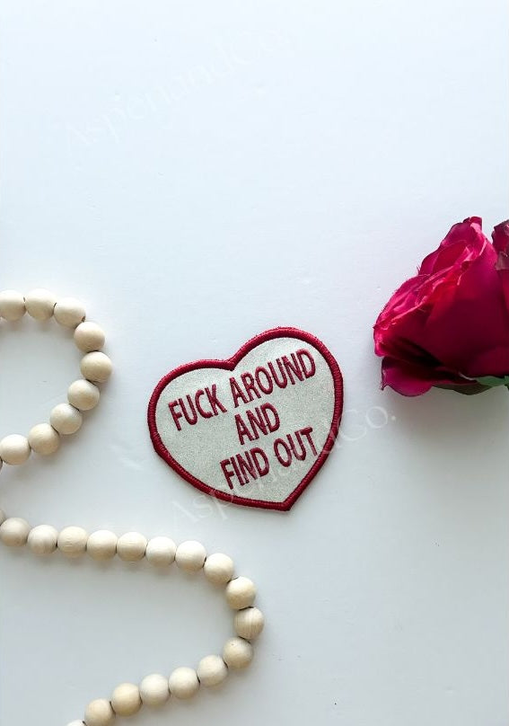 Glitter Heart-shaped patch with text 'F**K AROUND AND FIND OUT' on a white background with a pink rose and wooden beads.