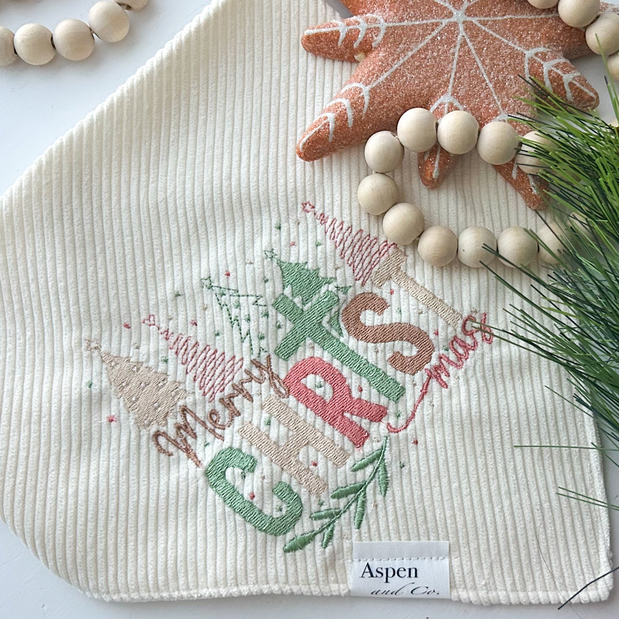 religious cream corduroy dog bandana embroidered with “Merry ChrisTmas” in pastel green, red, and gold thread, featuring Christmas trees and festive details. Snap-on handmade bandana by Aspen & Co., styled with holiday props close up.