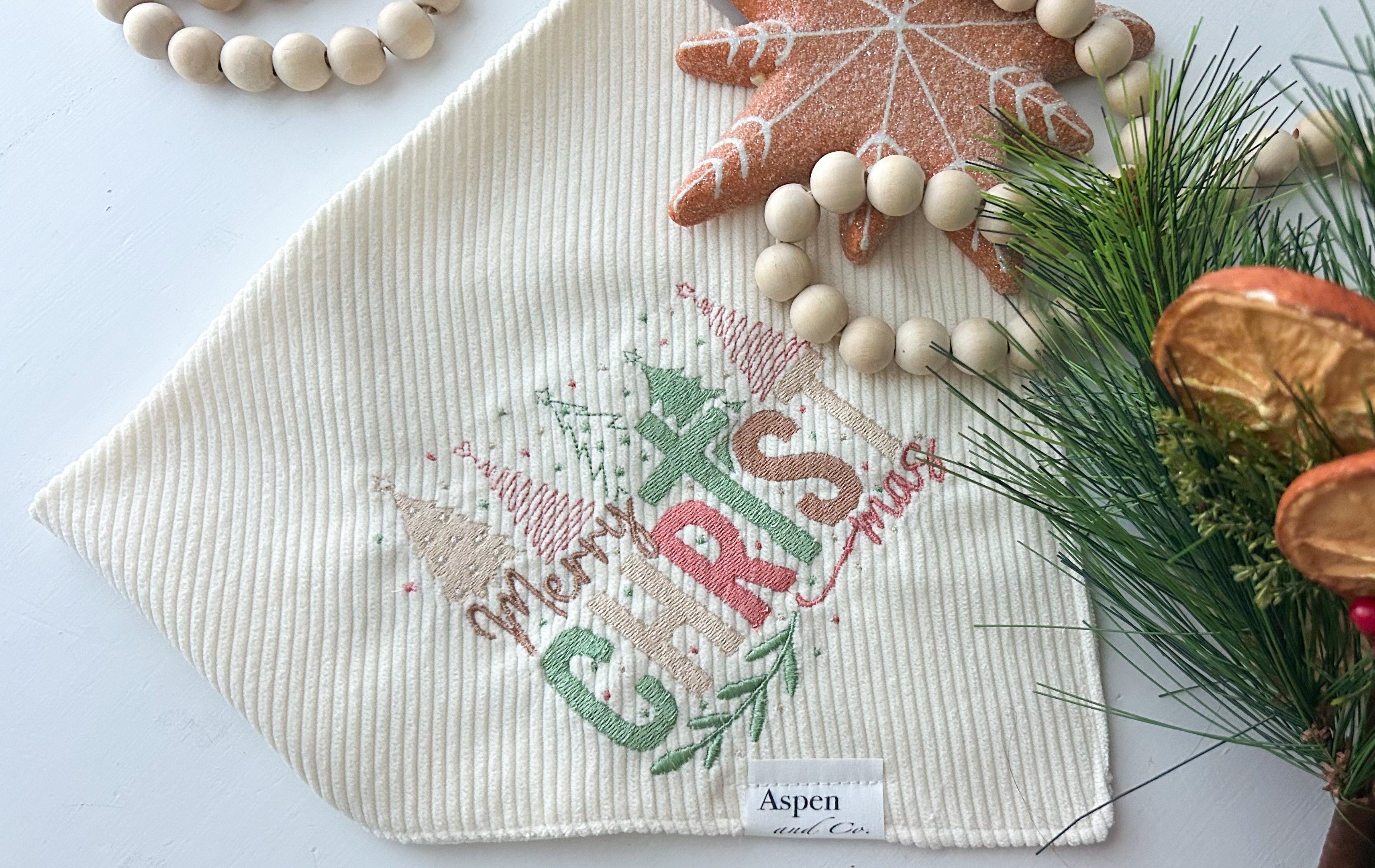 religious cream corduroy dog bandana embroidered with “Merry ChrisTmas” in pastel green, red, and gold thread, featuring Christmas trees and festive details. Snap-on handmade bandana by Aspen & Co., styled with holiday props close up.