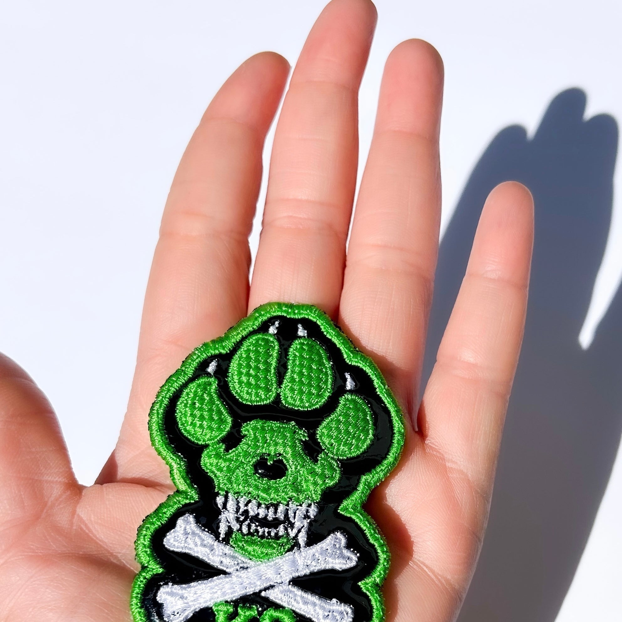 A hand holding a small embroidered patch shaped like a green and black paw print with a skull and crossbones design and the letters 'K9'. Text at the top says the patch is small, 3(1/8") tall x 2" wide, and advises double checking size suitability. Patch intended for dog harness, dog vest or backpack with loop side