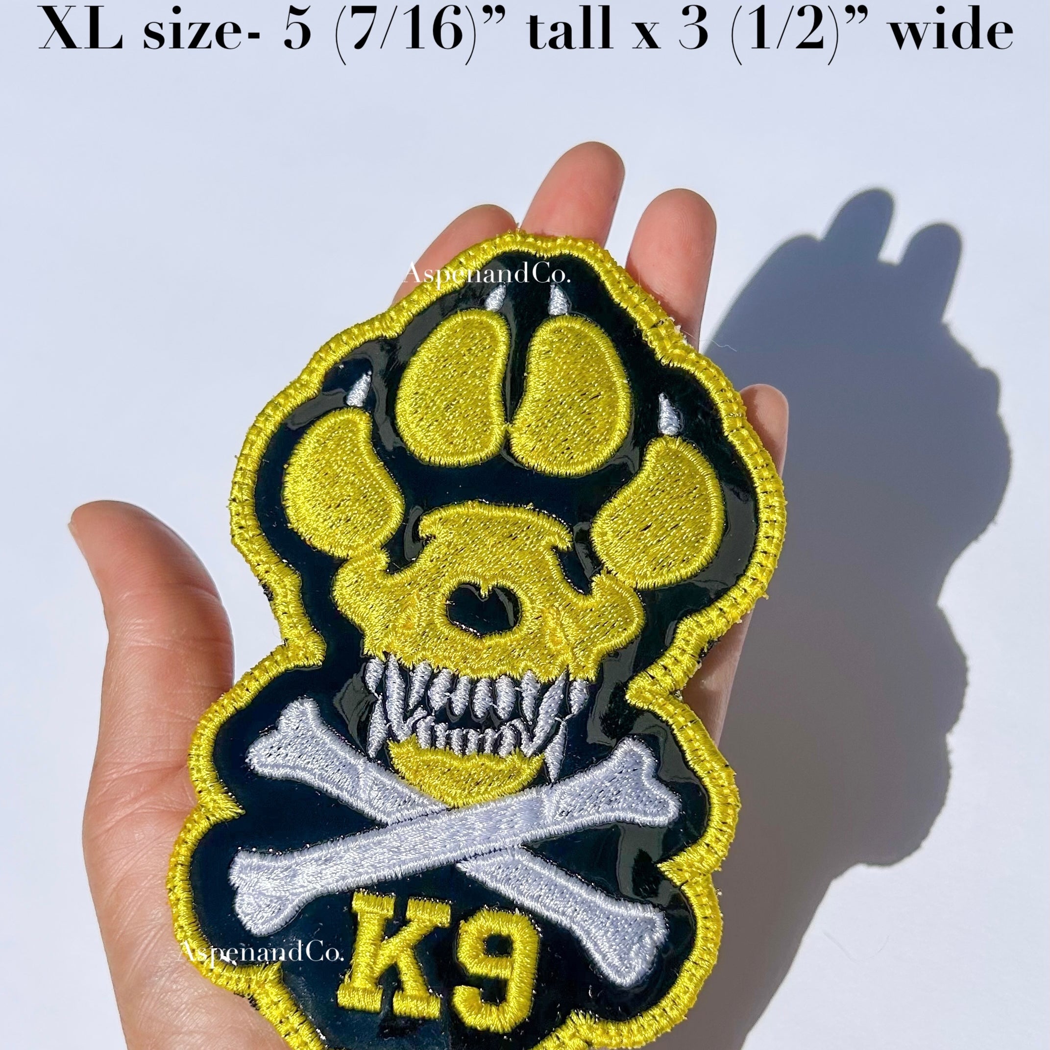 A hand holds a large embroidered patch shaped like a dog paw with a skull and crossbones, yellow 'K9' lettering, made in California by Aspen and Co. Text at the top reads 'XL size- 5 (7/16") tall x 3 (1/2") wide.