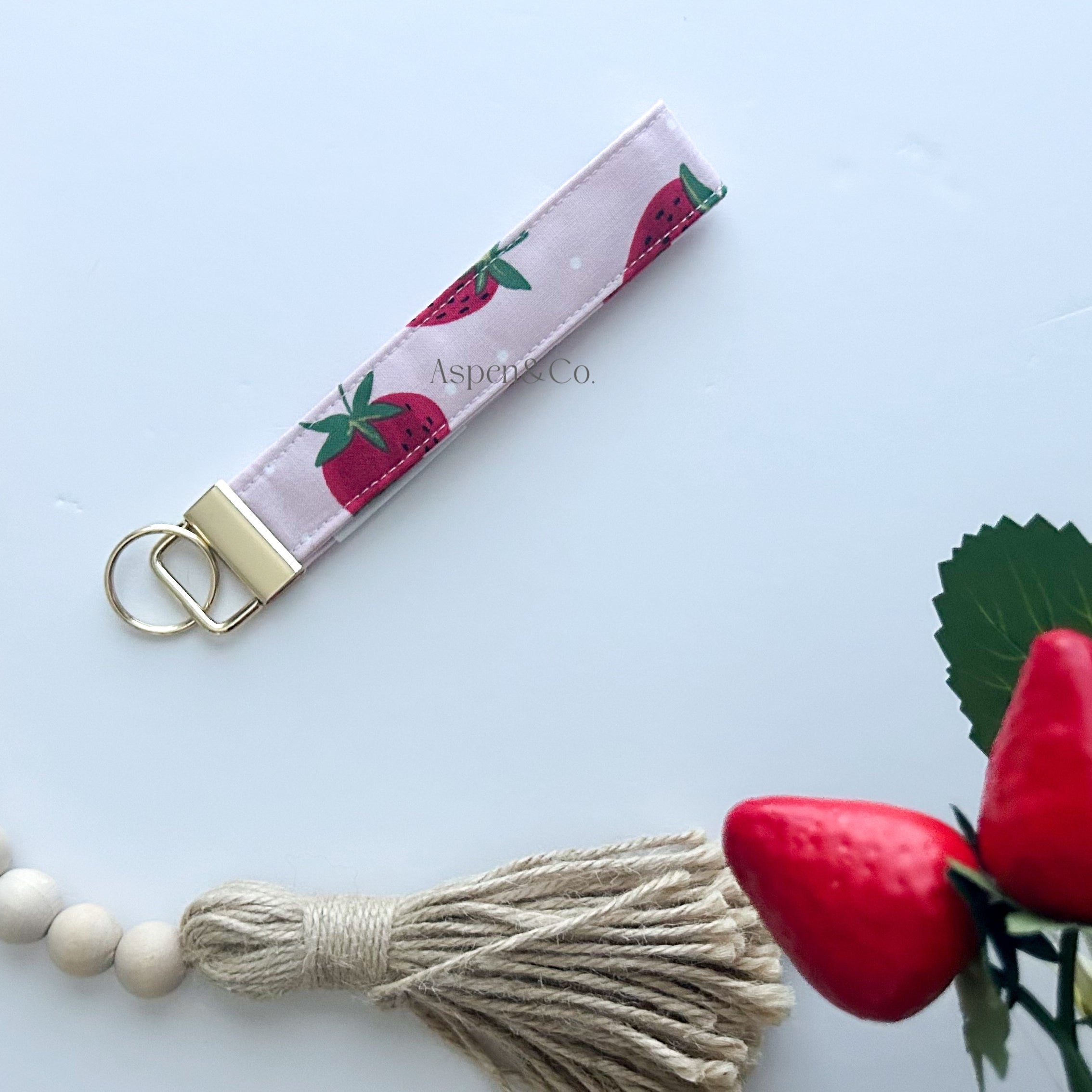 Wristlet keychain with strawberry pattern next to a wooden beaded necklace and red flowers on a light blue background