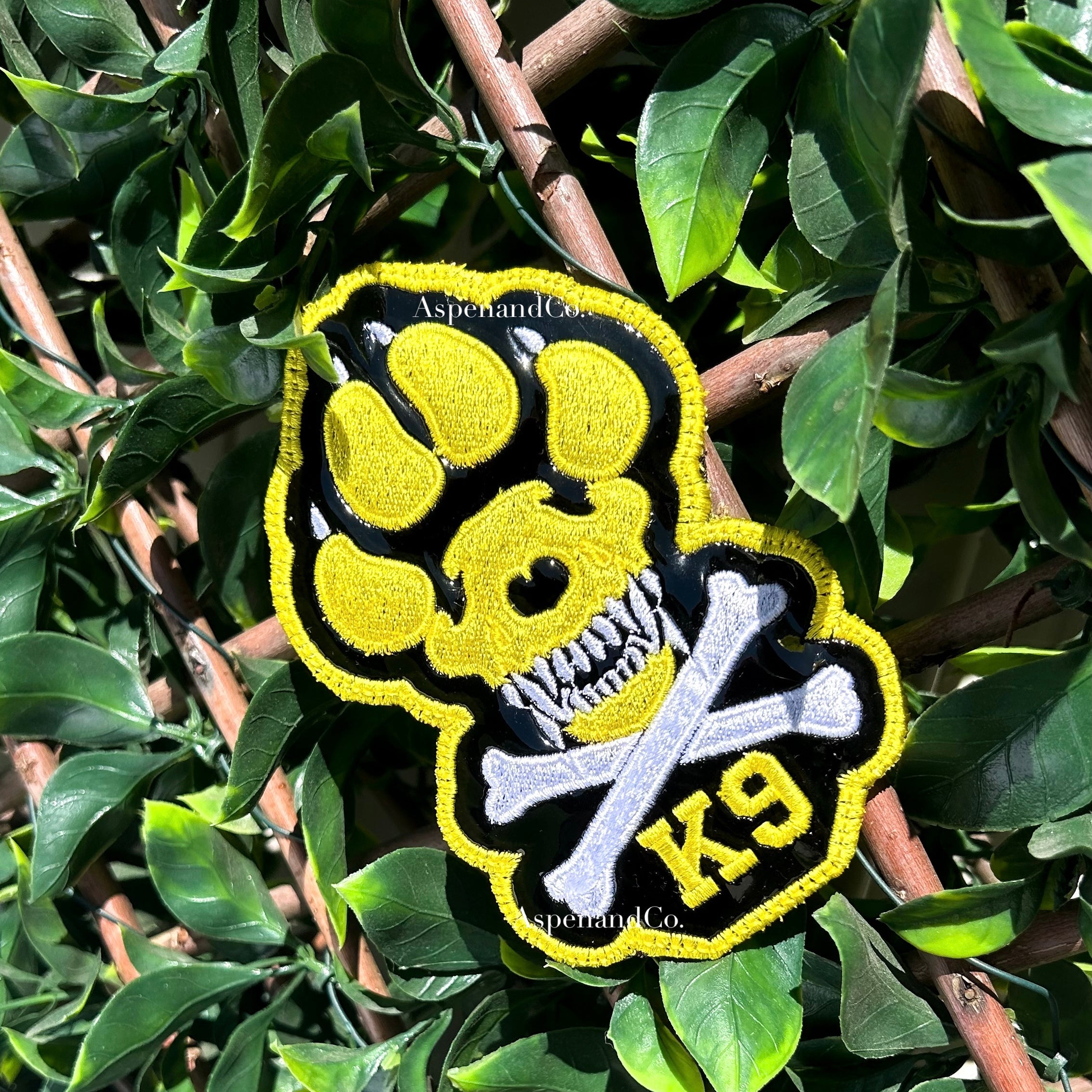 A yellow, black, and white embroidered K9 paw print patch with a skull and crossbones design, displayed on a green leafy branch.