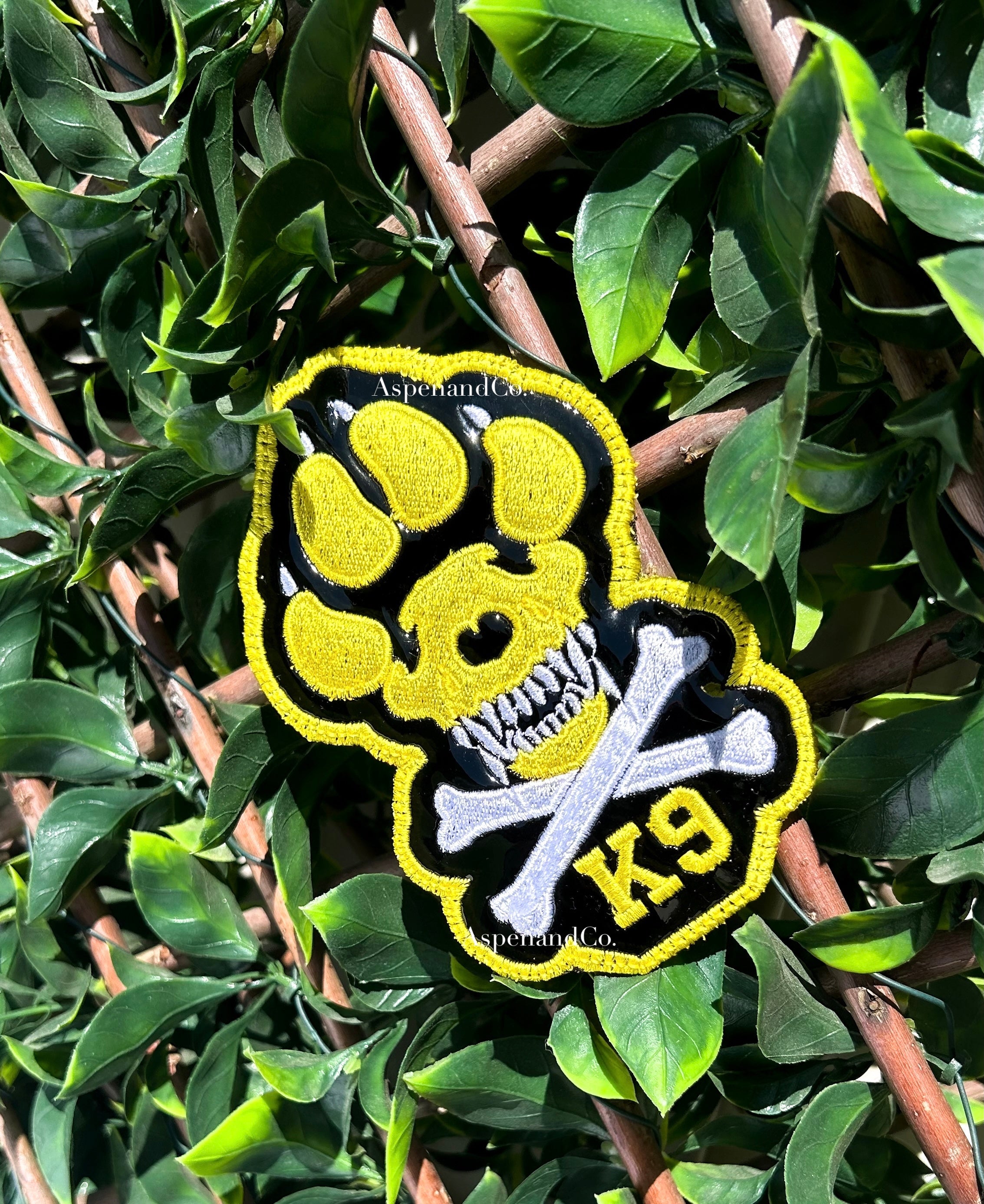 A yellow, black, and white embroidered K9 paw print patch with a skull and crossbones design, displayed on a green leafy branch.
