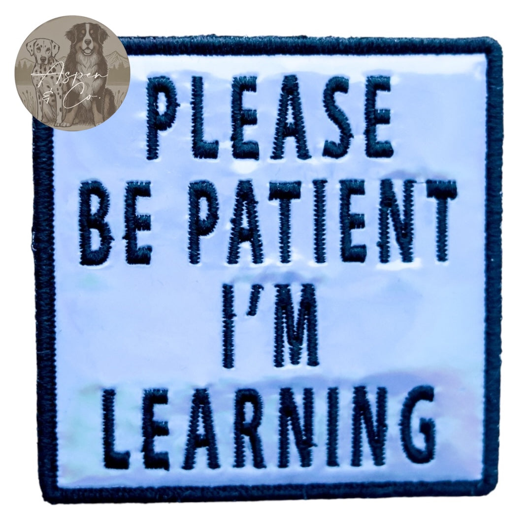 Blue holographic embroidered dog patch with black text that says 'Please Be Patient I'm Learning,' shown close-up with reflective detail. Patch can be attached to harnesses, capes, or vests.