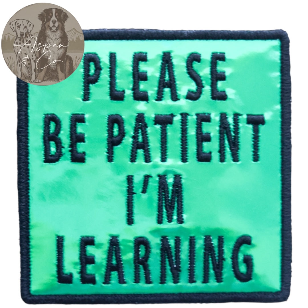 Green holographic embroidered dog patch with black text reading 'Please Be Patient I'm Learning,' shown close-up highlighting reflective finish and stitching. Suitable for attaching to dog harnesses, capes, or vests.