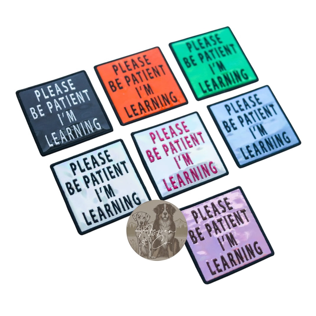 black, orange, green, silver with black text, silver with pink text, blue, and pink, all embroidered with “Be Patient I’m Learning.”