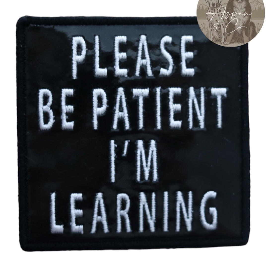 Embroidered black holographic dog patch with white text that reads 'Please Be Patient I'm Learning,' shown close-up to highlight shine and stitching detail.
