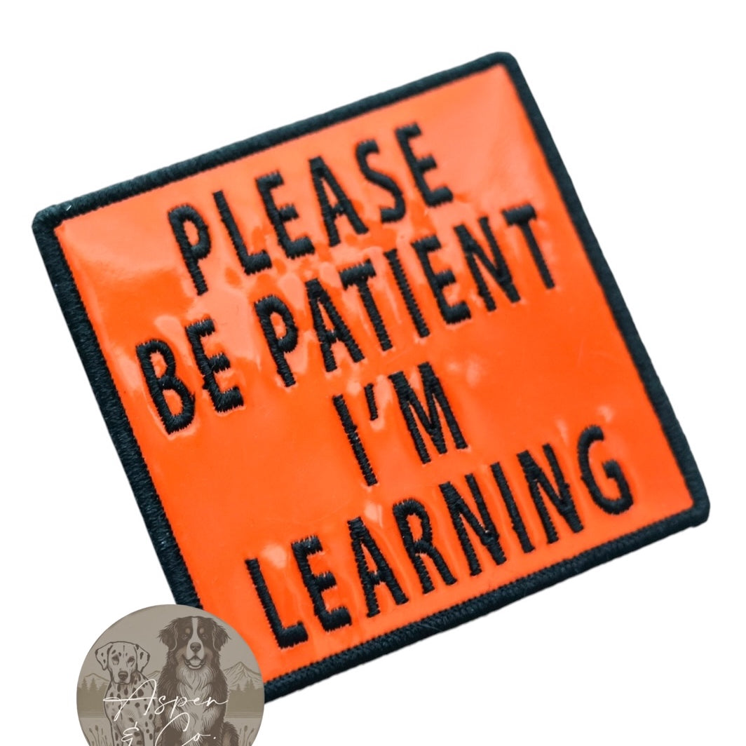 Orange holographic embroidered dog patch with black text reading 'Please Be Patient I'm Learning,' shown close-up with reflective shine and detailed stitching. Perfect for attaching to dog harnesses, capes, or vests.