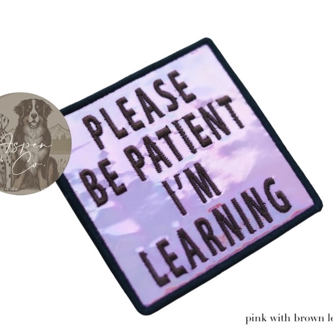 Pink holographic embroidered dog patch with brown text reading 'Please Be Patient I'm Learning,' shown close-up highlighting reflective finish and stitching detail. Designed to attach to dog harnesses, capes, or vests.