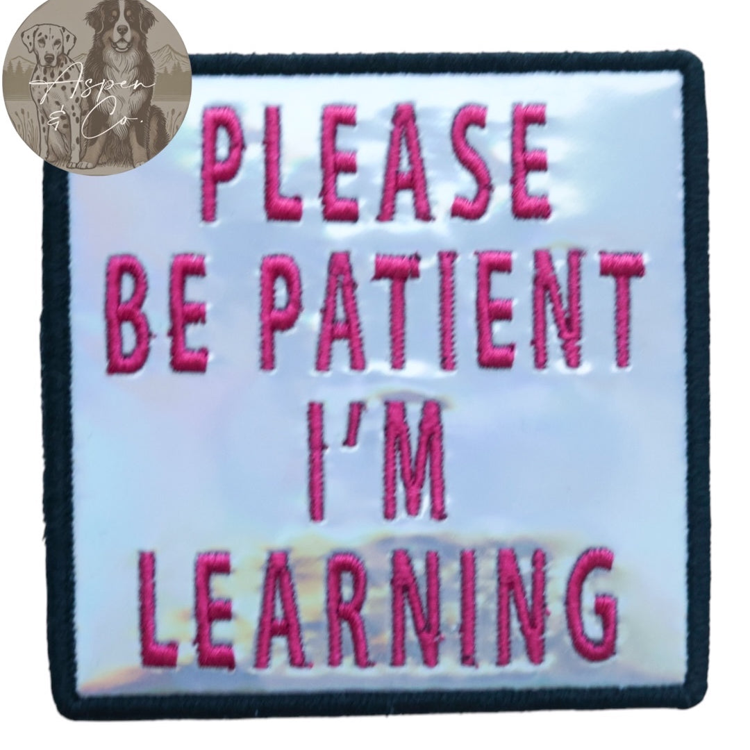 Silver holographic embroidered dog patch with hot pink text reading 'Please Be Patient I'm Learning,' shown close-up highlighting shiny reflective finish and detailed stitching. Ideal for attaching to dog harnesses, capes, or vests.