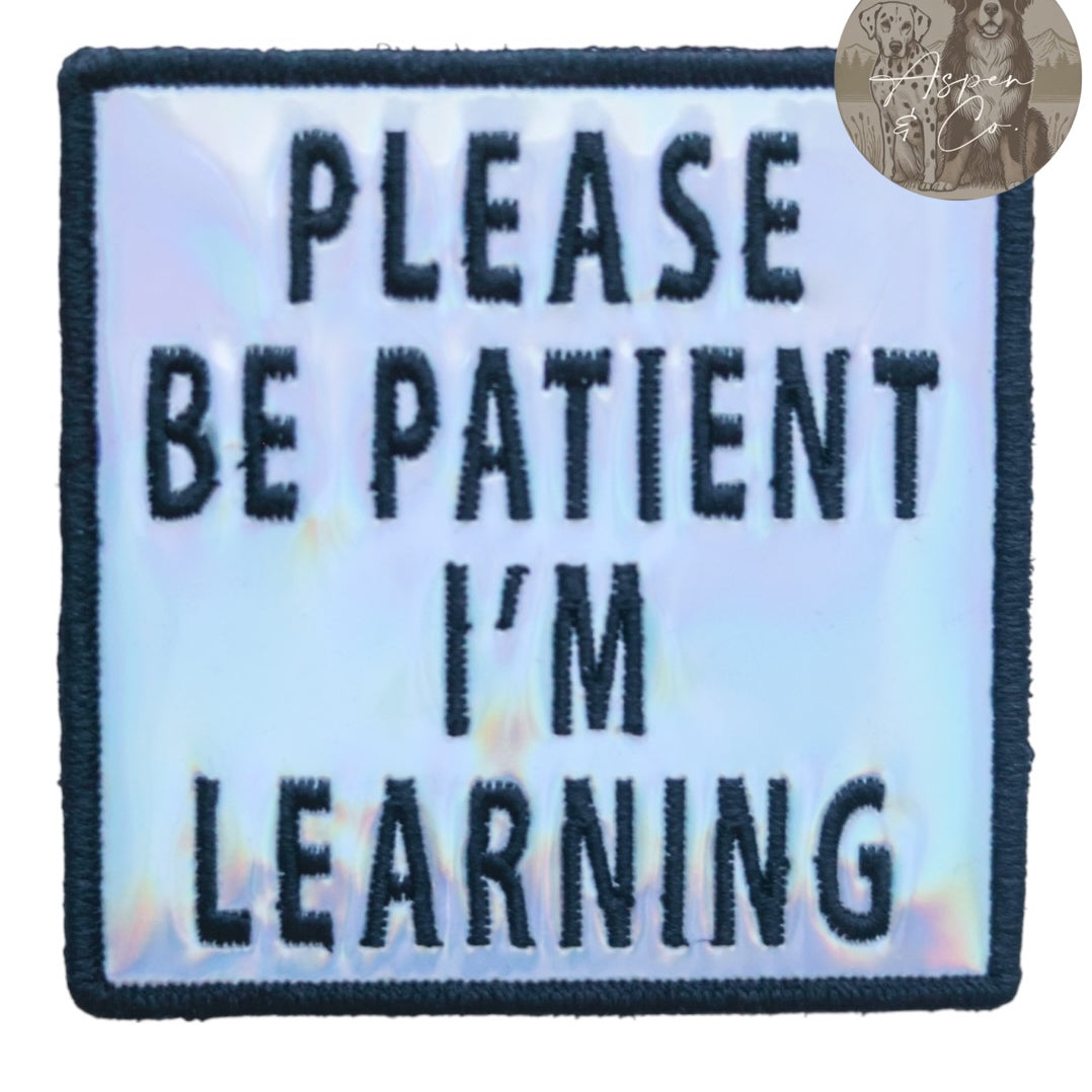 Silver holographic embroidered dog patch with black text saying 'Please Be Patient I'm Learning,' shown close-up with shiny reflective detail. Designed to attach easily to dog harnesses, capes, or vests.