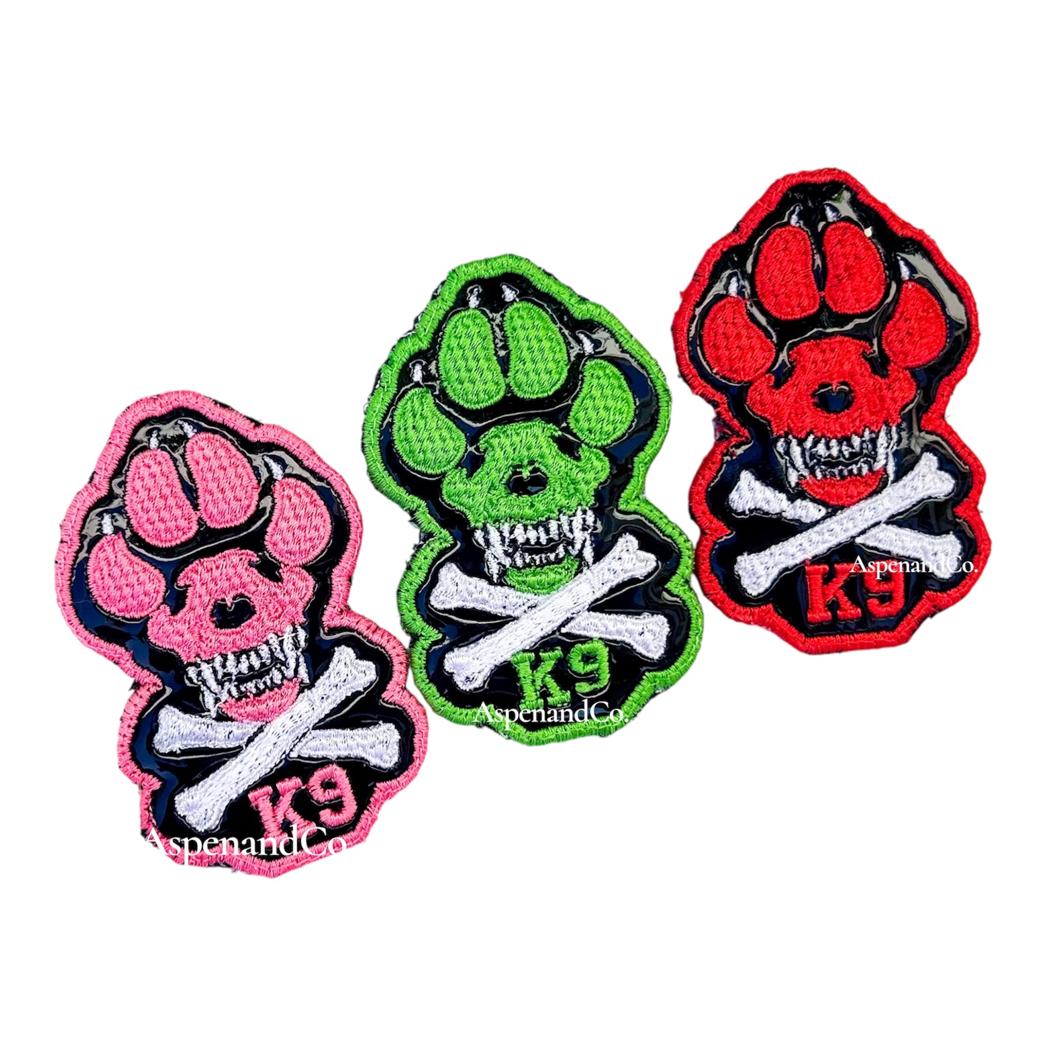 Three embroidered dog paw patch designs with crossed bones and skull motifs, shown on a plain white background. The patches are pink, green, and red, each featuring the text ' K9.' Made by 'Aspen and Co' in California