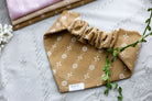 Back view of the ochre floral dog bandana with scrunchie neckline, displayed on a sandy background alongside white flowers and coordinating woven fabric options.