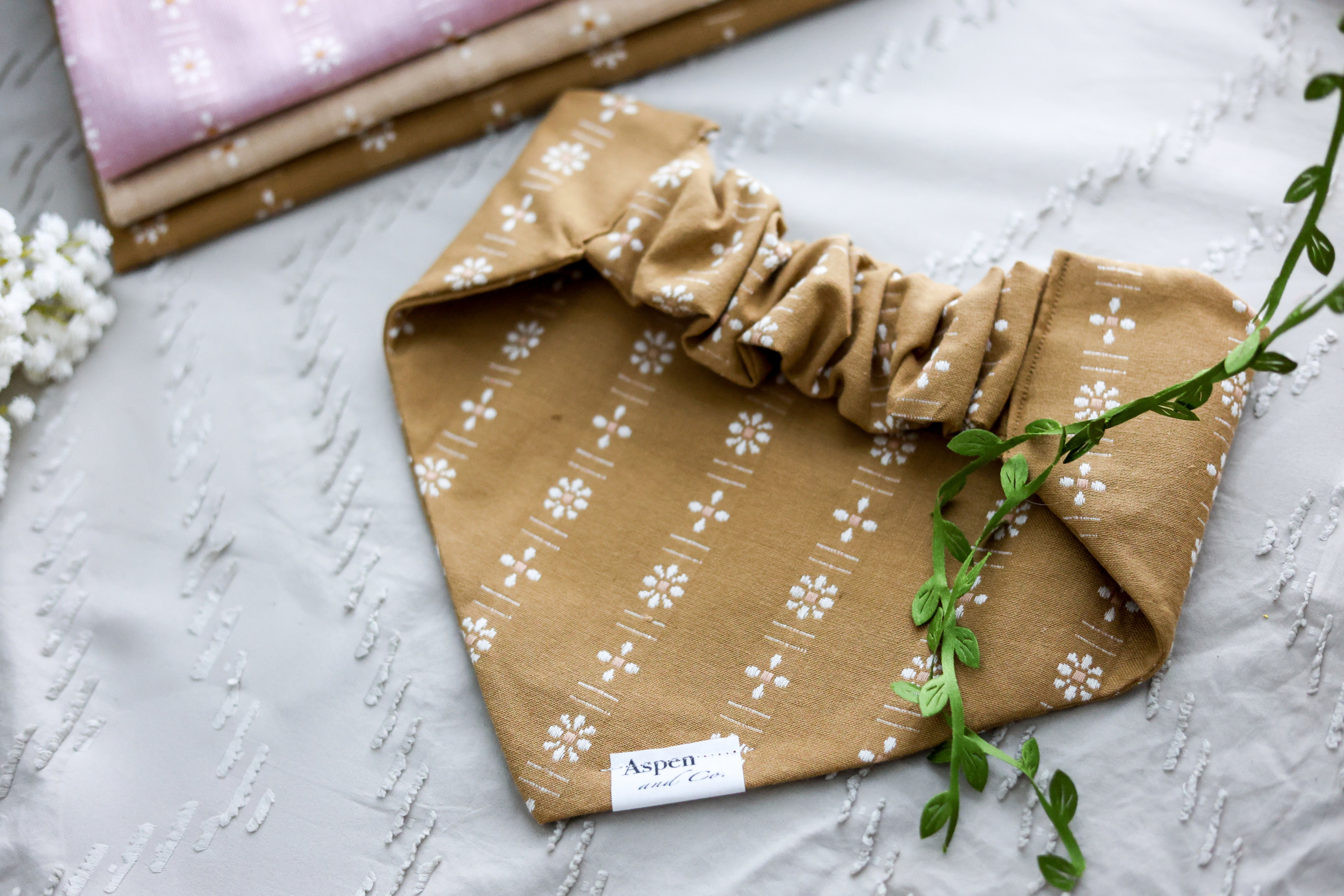 Back view of the ochre floral dog bandana with scrunchie neckline, displayed on a sandy background alongside white flowers and coordinating woven fabric options.