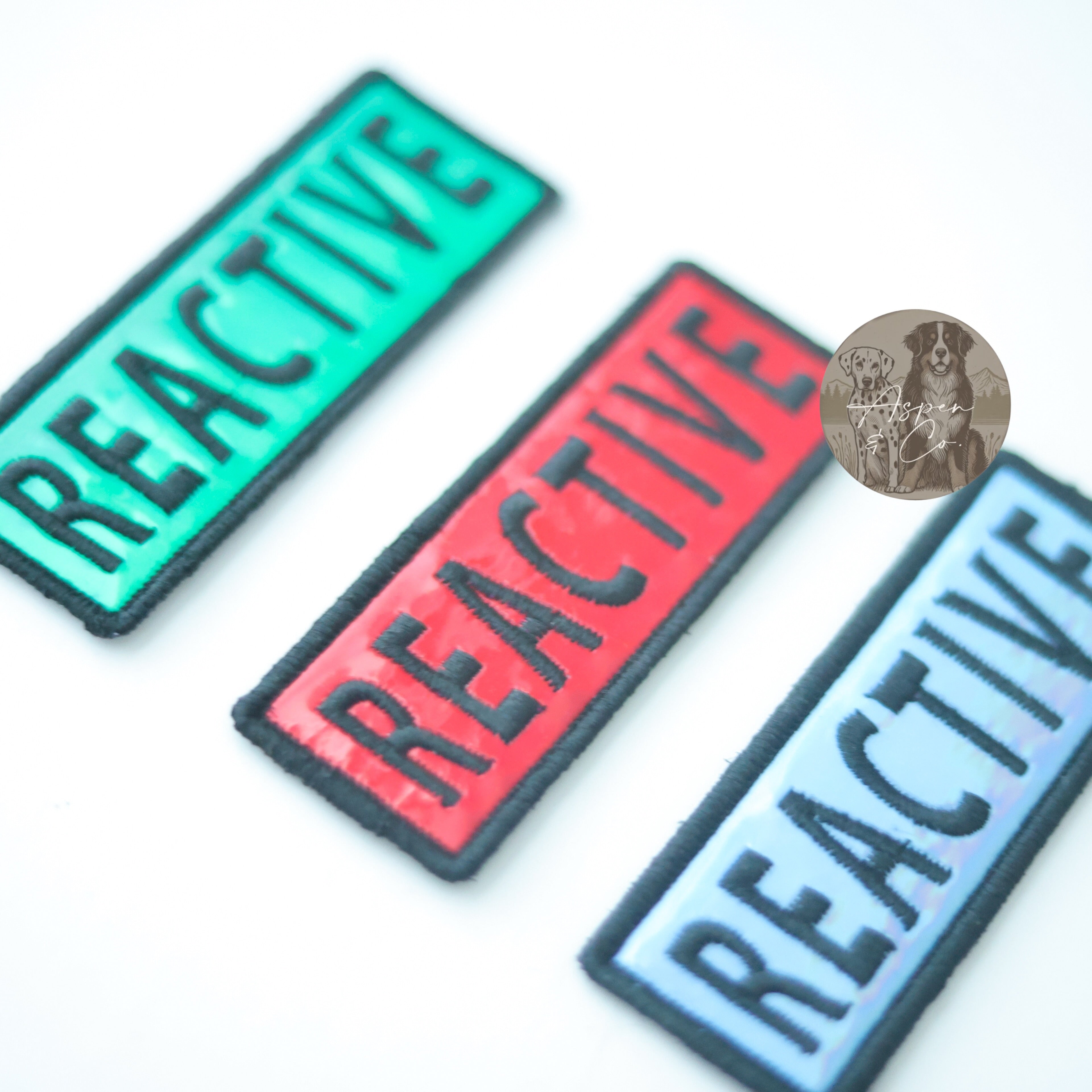 Photo displaying holographic dog patches with the word "Reactive" in multiple color options, perfect for attaching to collars, harnesses, vests, or capes.