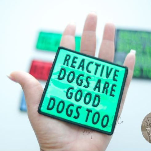 Close-up photo of a greenk holographic dog patch with neon green text reading "Reactive Dogs Are Good Dogs Too," designed for harnesses, vests, or capes.