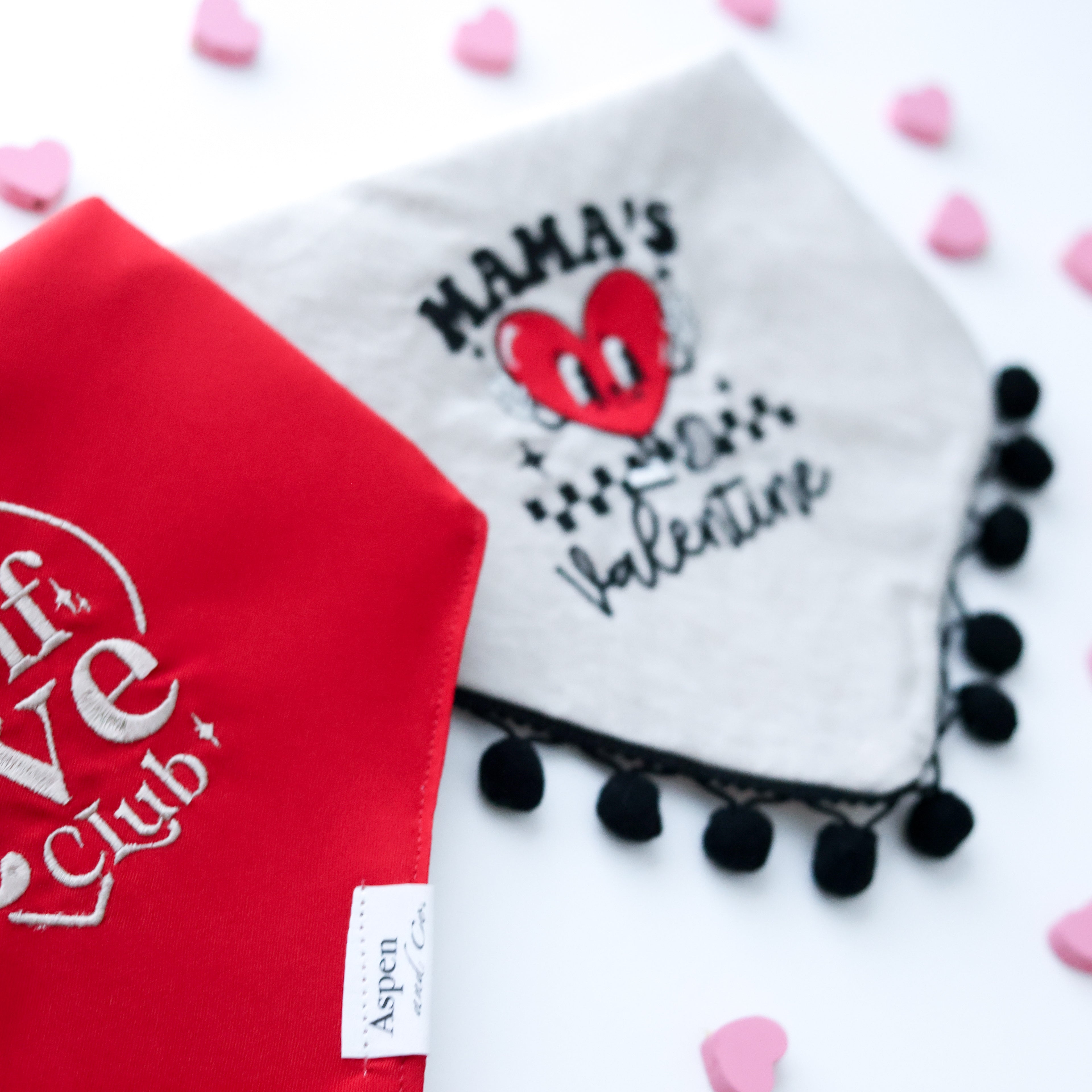 Two embroidered dog bandanas side by side—one cool-toned red bandana with beige stitching that says “Self Love Club” inside a heart, and one pink linen bandana with red embroidery reading “Mama’s Valentine”; both are stylish Valentine’s Day accessories for dogs.