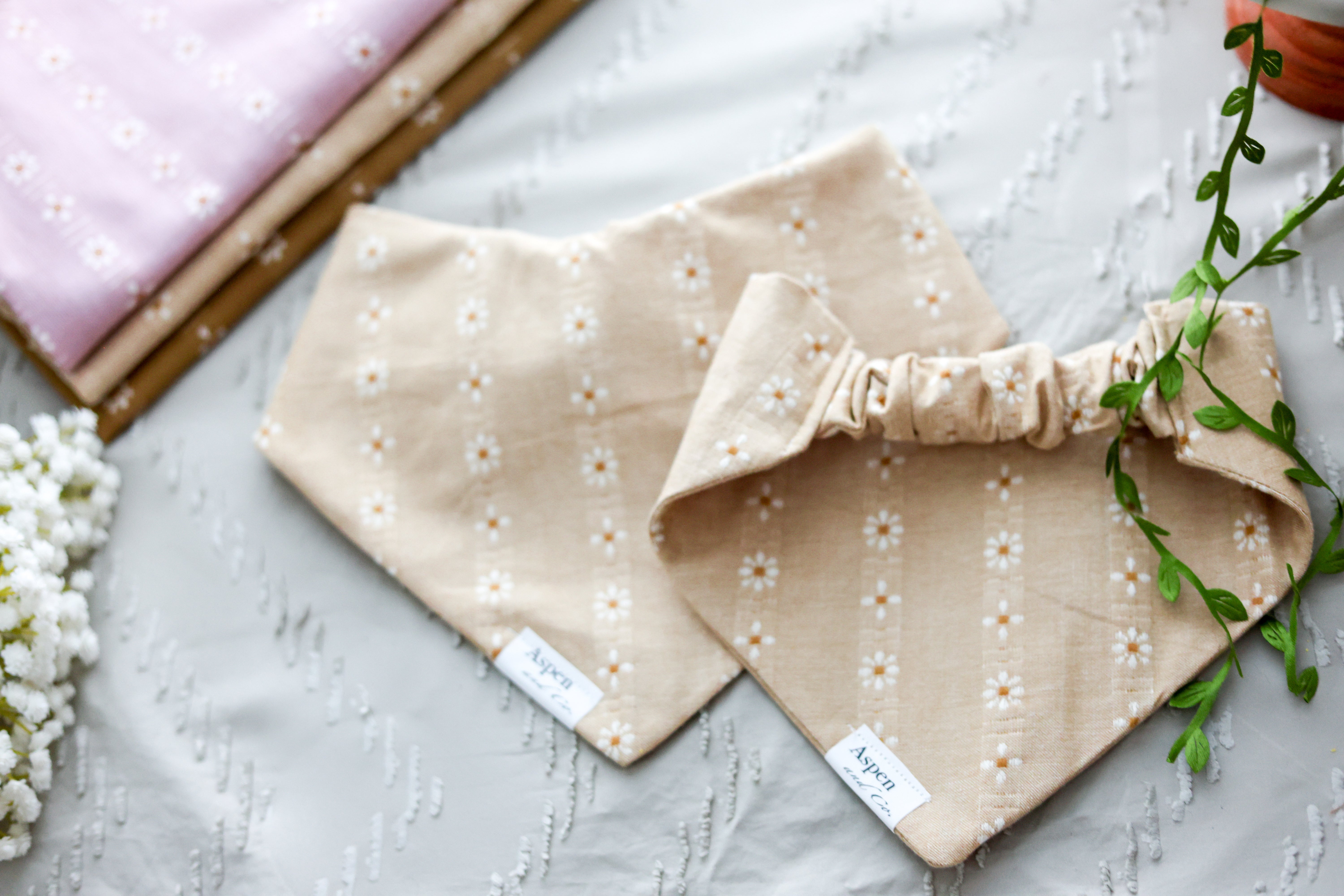 Flat lay of camel floral dog bandana shown front and back on a sandy background with white flowers and other woven fabric color options