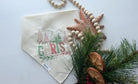 Cream corduroy dog bandana embroidered with “Merry ChrisTmas” in pastel green, red, and gold thread, featuring Christmas trees and festive details. Snap-on handmade bandana by Aspen & Co., styled with holiday props.