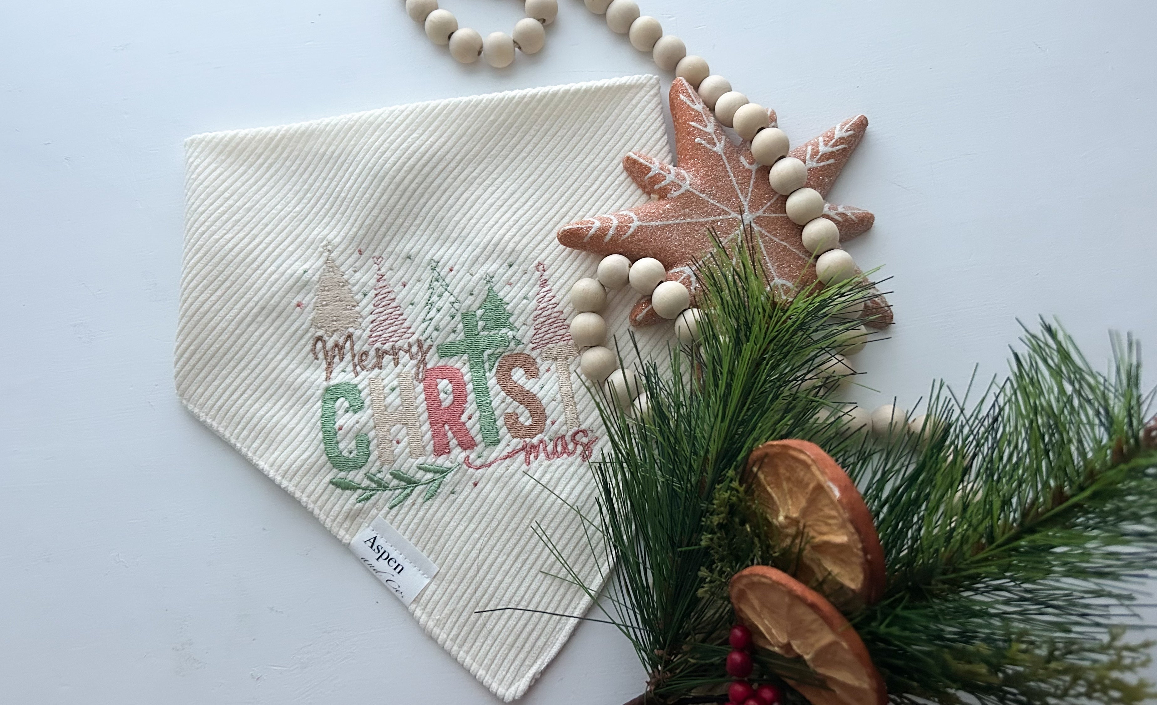 Cream corduroy dog bandana embroidered with “Merry ChrisTmas” in pastel green, red, and gold thread, featuring Christmas trees and festive details. Snap-on handmade bandana by Aspen & Co., styled with holiday props.