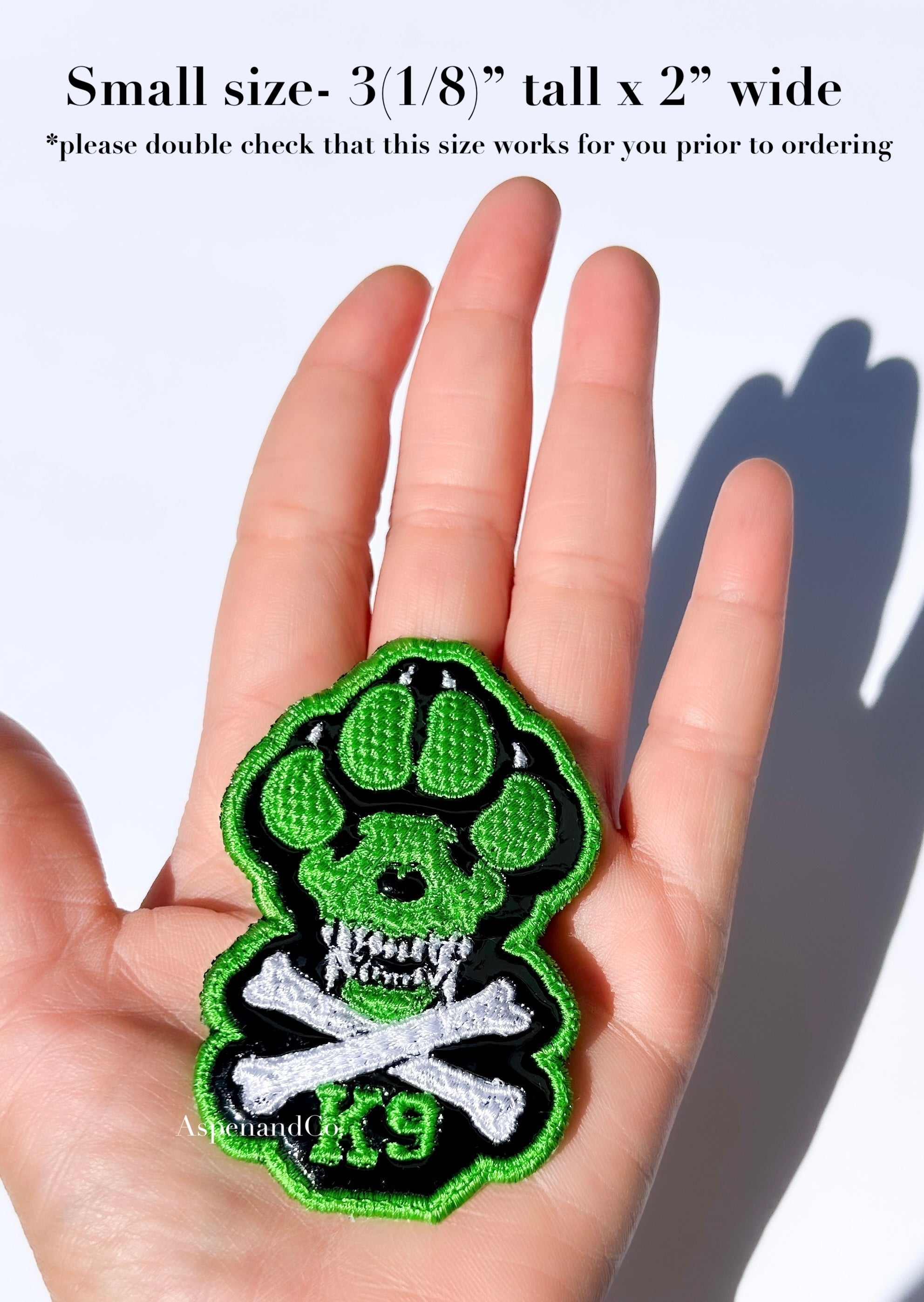A hand holding a small embroidered patch shaped like a green and black paw print with a skull and crossbones design and the letters 'K9'. Text at the top says the patch is small, 3(1/8") tall x 2" wide, and advises double checking size suitability. Patch intended for dog harness, dog vest or backpack with loop side
