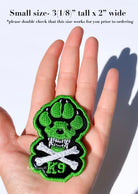 A hand holding a small embroidered patch shaped like a green and black paw print with a skull and crossbones design and the letters 'K9'. Text at the top says the patch is small, 3(1/8") tall x 2" wide, and advises double checking size suitability. Patch intended for dog harness, dog vest or backpack with loop side