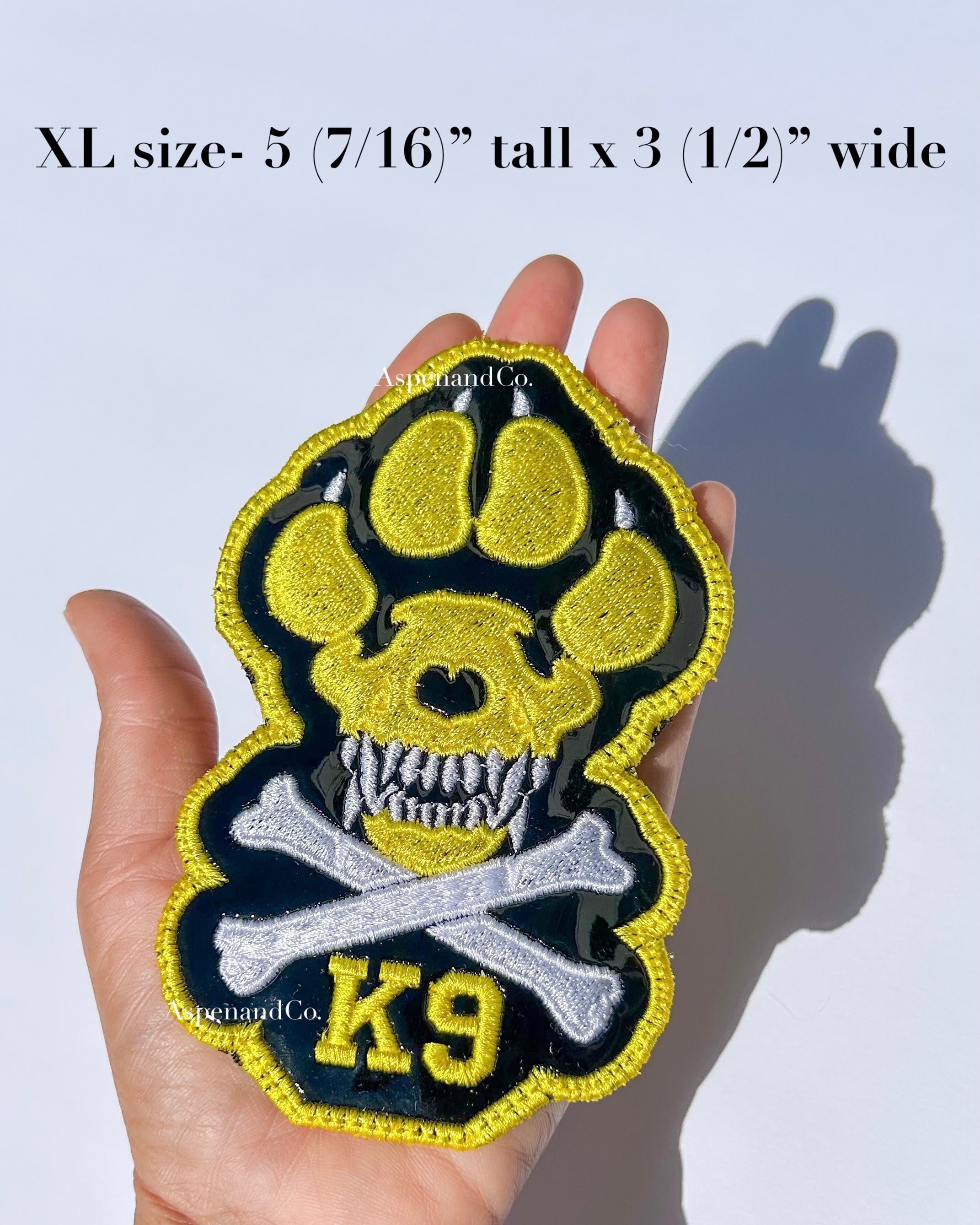 A hand holds a large embroidered patch shaped like a dog paw with a skull and crossbones, yellow 'K9' lettering, made in California by Aspen and Co. Text at the top reads 'XL size- 5 (7/16") tall x 3 (1/2") wide.