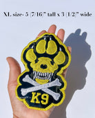 A hand holds a large embroidered patch shaped like a dog paw with a skull and crossbones, yellow 'K9' lettering, made in California by Aspen and Co. Text at the top reads 'XL size- 5 (7/16") tall x 3 (1/2") wide.