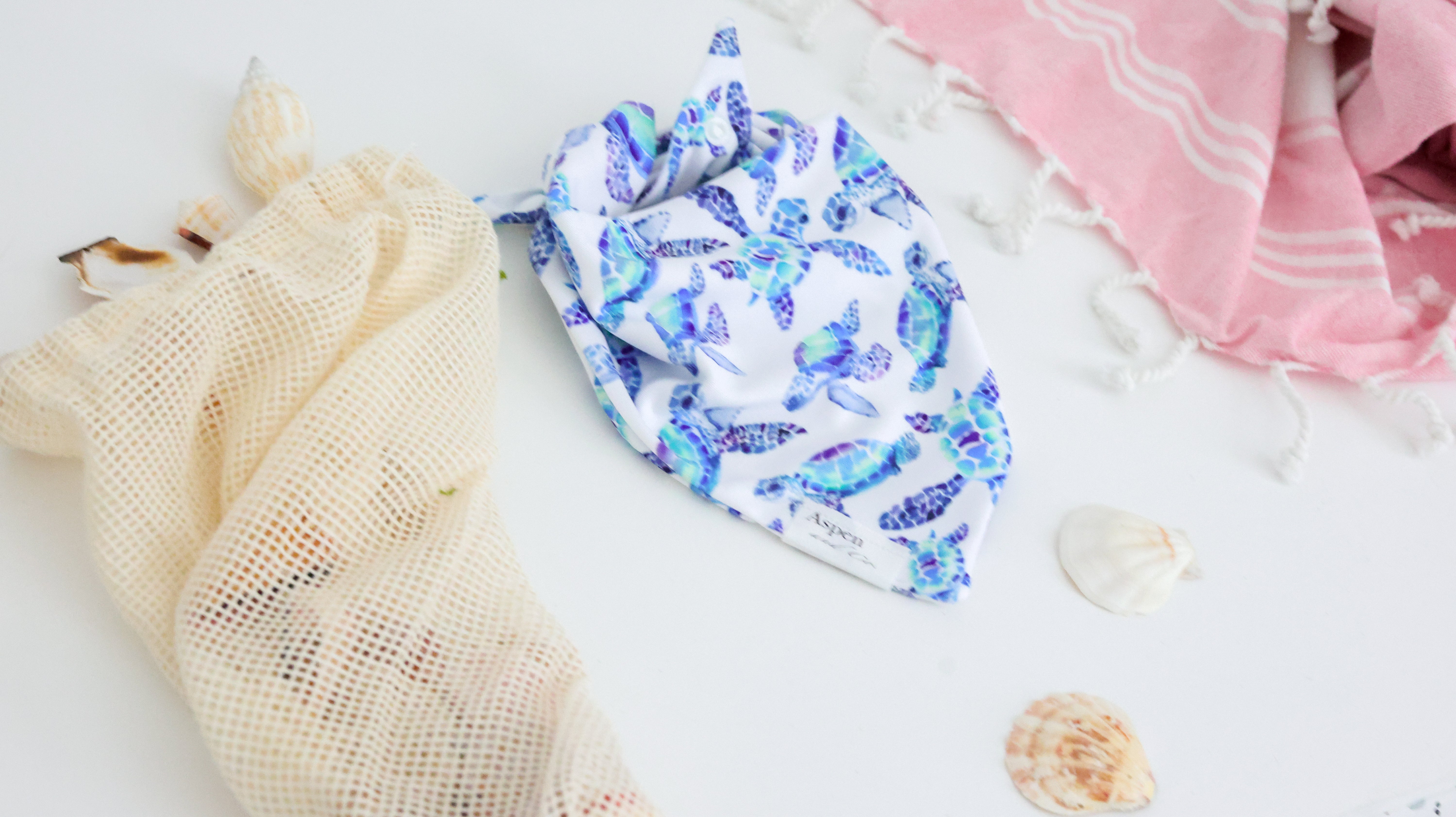 White weatherproof dog bandana with blue and turquoise sea turtles, Aspen and Co. tag, styled with seashells and a beach towel