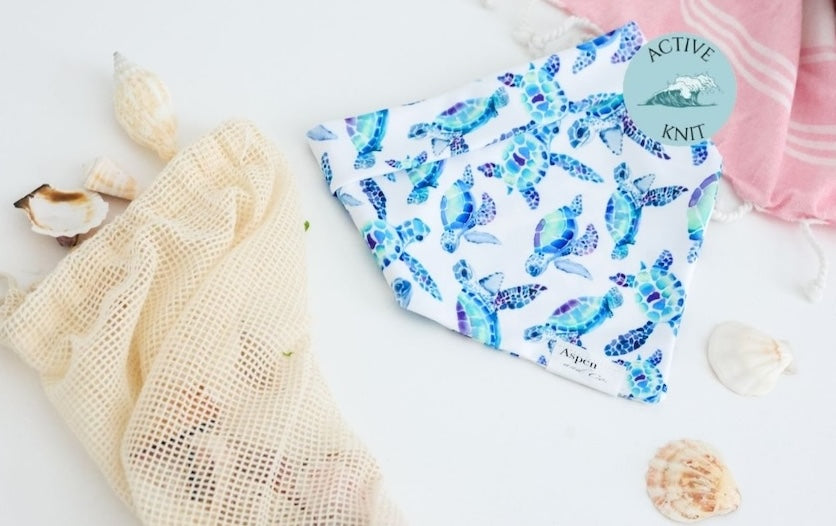 White weatherproof dog bandana with blue and turquoise sea turtles, Aspen and Co. tag, styled with seashells and a beach towel