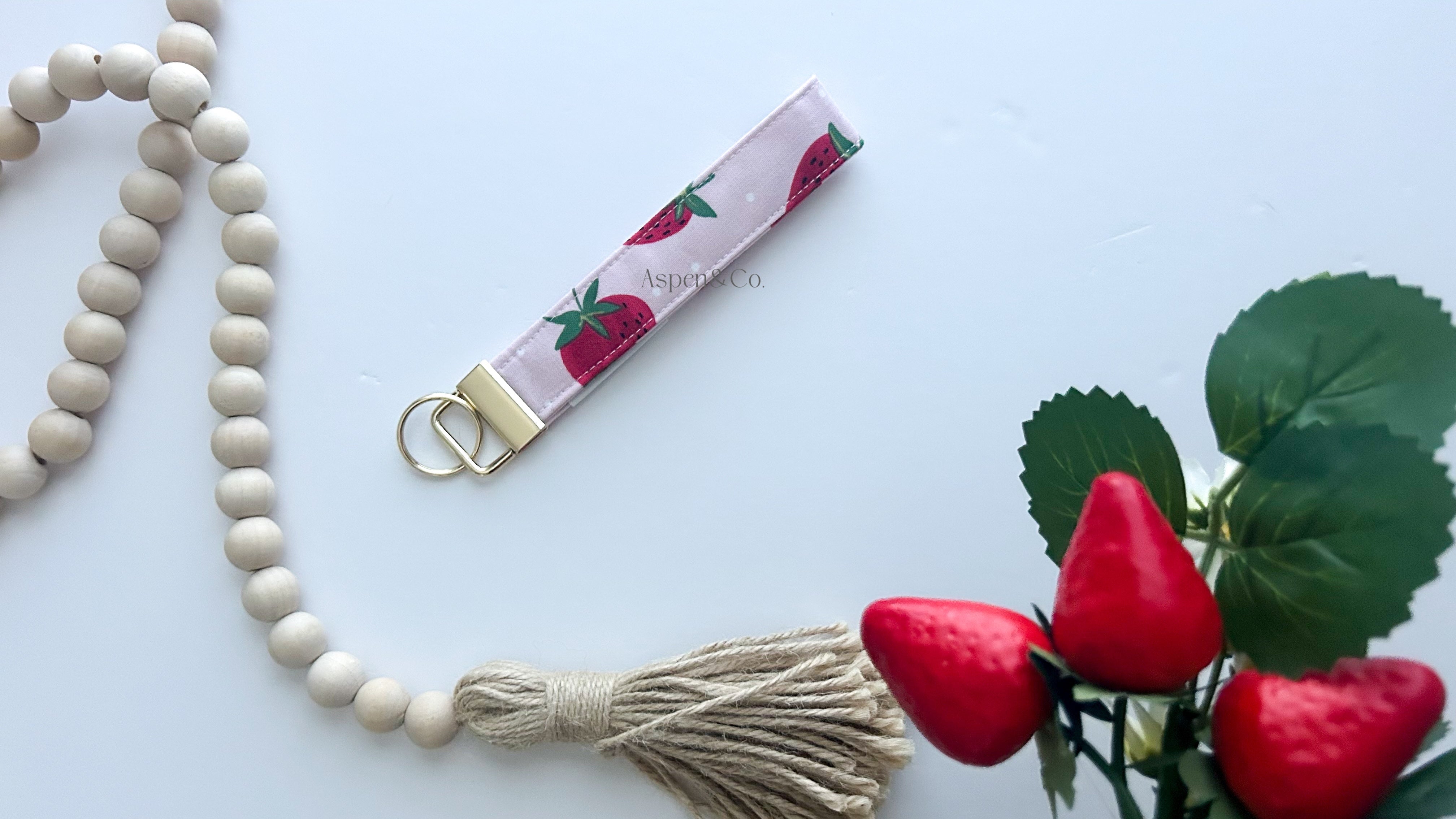 Wristlet keychain with strawberry pattern next to a wooden beaded necklace and red flowers on a light blue background