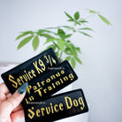 Three black patches with gold text held by a hand, with a plant in the background.