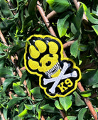A yellow, black, and white embroidered K9 paw print patch with a skull and crossbones design, displayed on a green leafy branch.