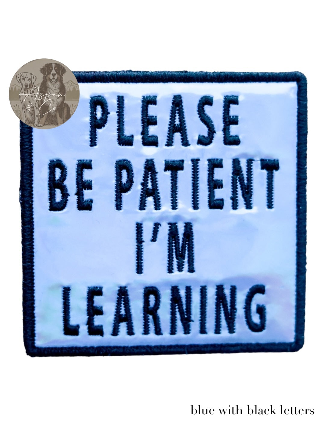 Blue holographic embroidered dog patch with black text that says 'Please Be Patient I'm Learning,' shown close-up with reflective detail. Patch can be attached to harnesses, capes, or vests.