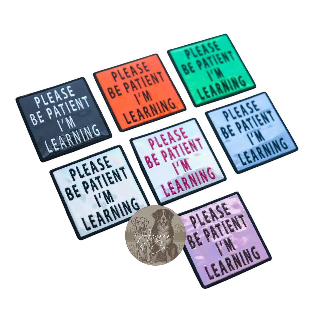 black, orange, green, silver with black text, silver with pink text, blue, and pink, all embroidered with “Be Patient I’m Learning.”