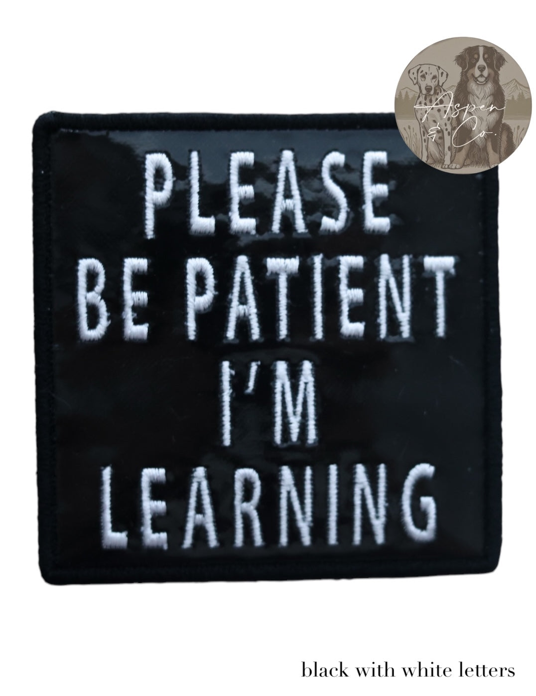 Embroidered black holographic dog patch with white text that reads 'Please Be Patient I'm Learning,' shown close-up to highlight shine and stitching detail.
