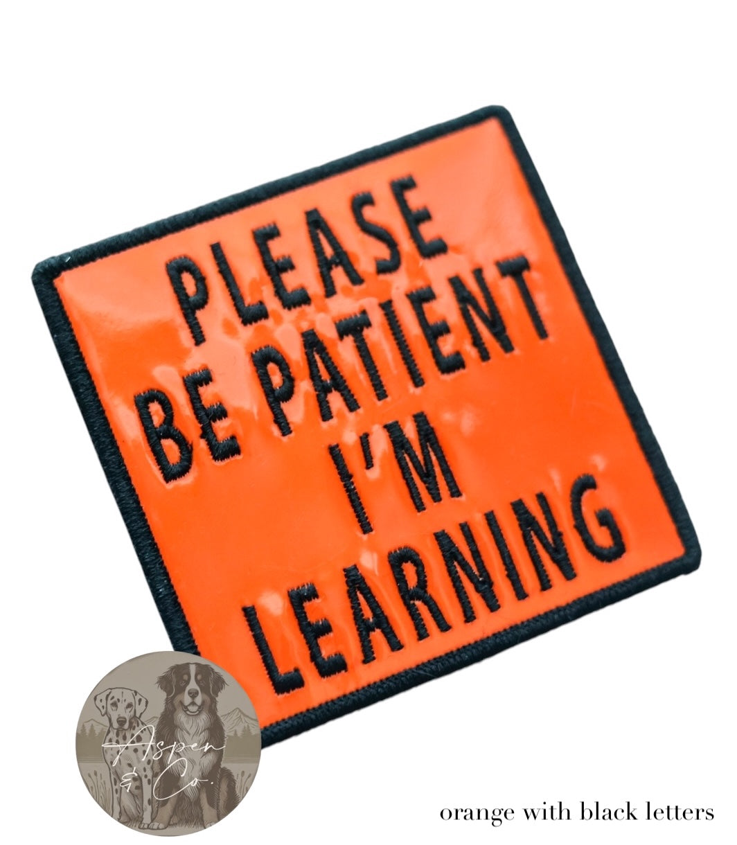 Orange holographic embroidered dog patch with black text reading 'Please Be Patient I'm Learning,' shown close-up with reflective shine and detailed stitching. Perfect for attaching to dog harnesses, capes, or vests.