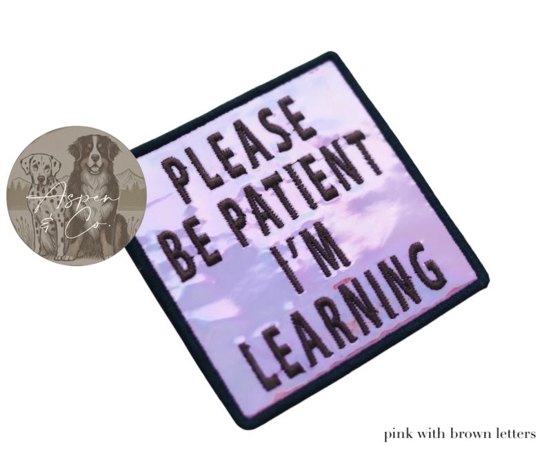 Pink holographic embroidered dog patch with brown text reading 'Please Be Patient I'm Learning,' shown close-up highlighting reflective finish and stitching detail. Designed to attach to dog harnesses, capes, or vests.