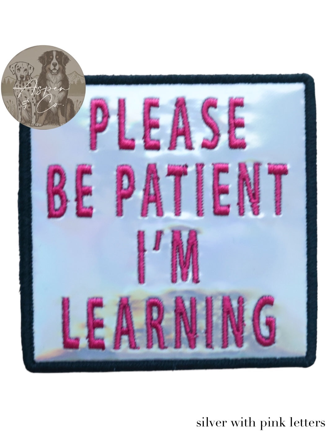 Silver holographic embroidered dog patch with hot pink text reading 'Please Be Patient I'm Learning,' shown close-up highlighting shiny reflective finish and detailed stitching. Ideal for attaching to dog harnesses, capes, or vests.