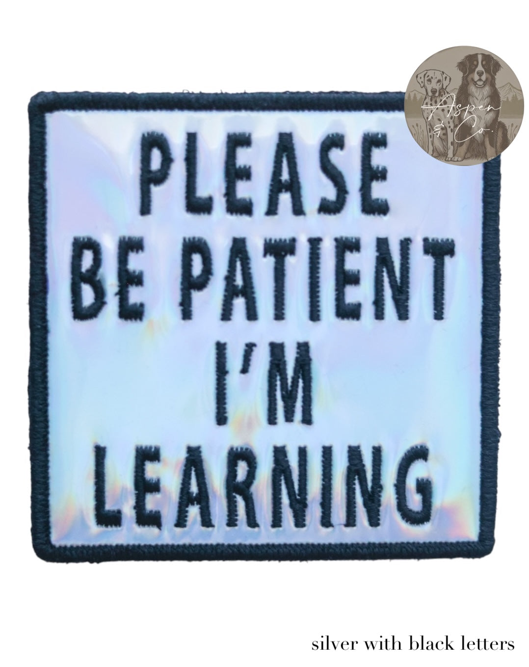 Silver holographic embroidered dog patch with black text saying 'Please Be Patient I'm Learning,' shown close-up with shiny reflective detail. Designed to attach easily to dog harnesses, capes, or vests.