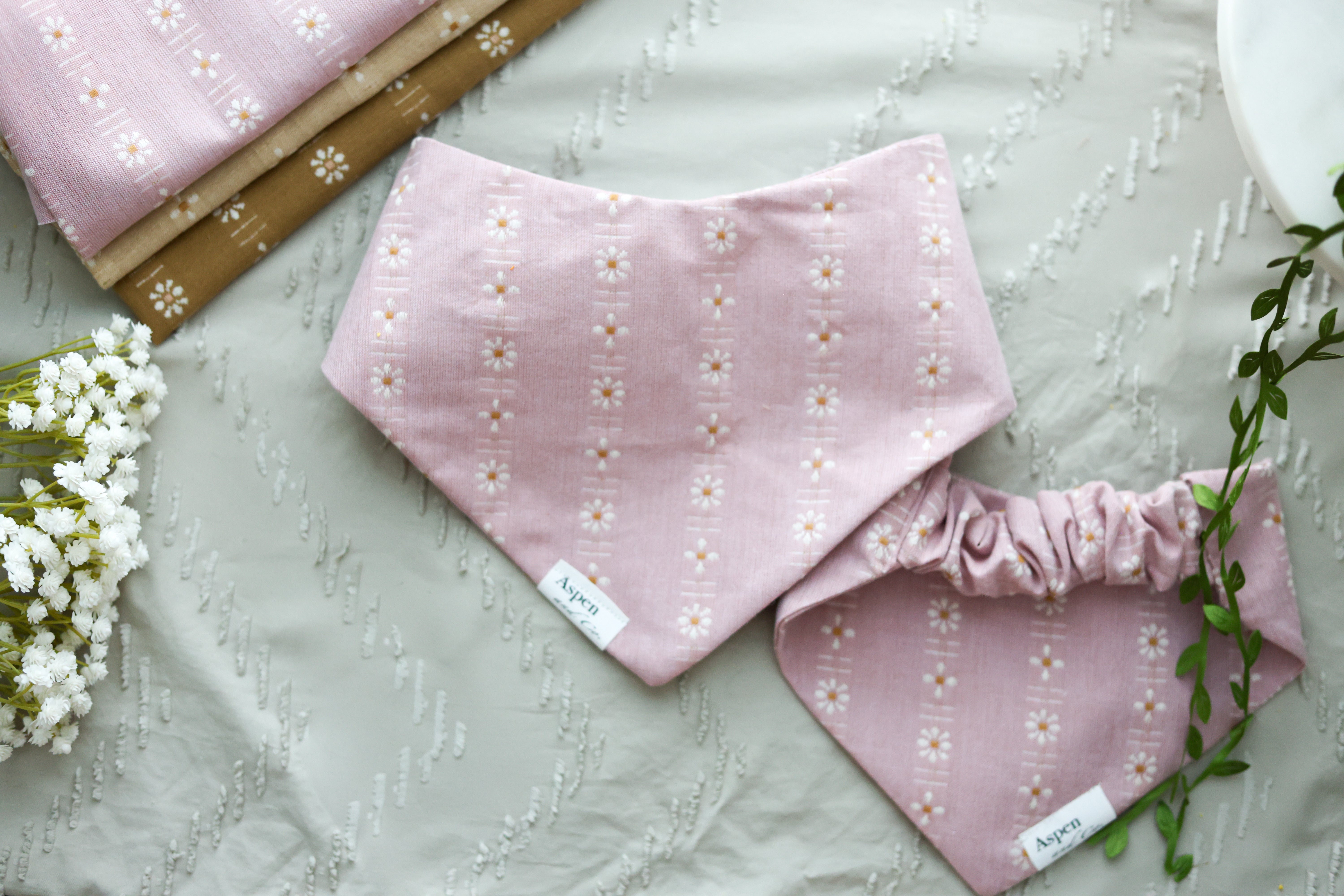 Flat lay of pink floral dog bandana shown front and back on a sandy background with white flowers and other woven fabric color options.