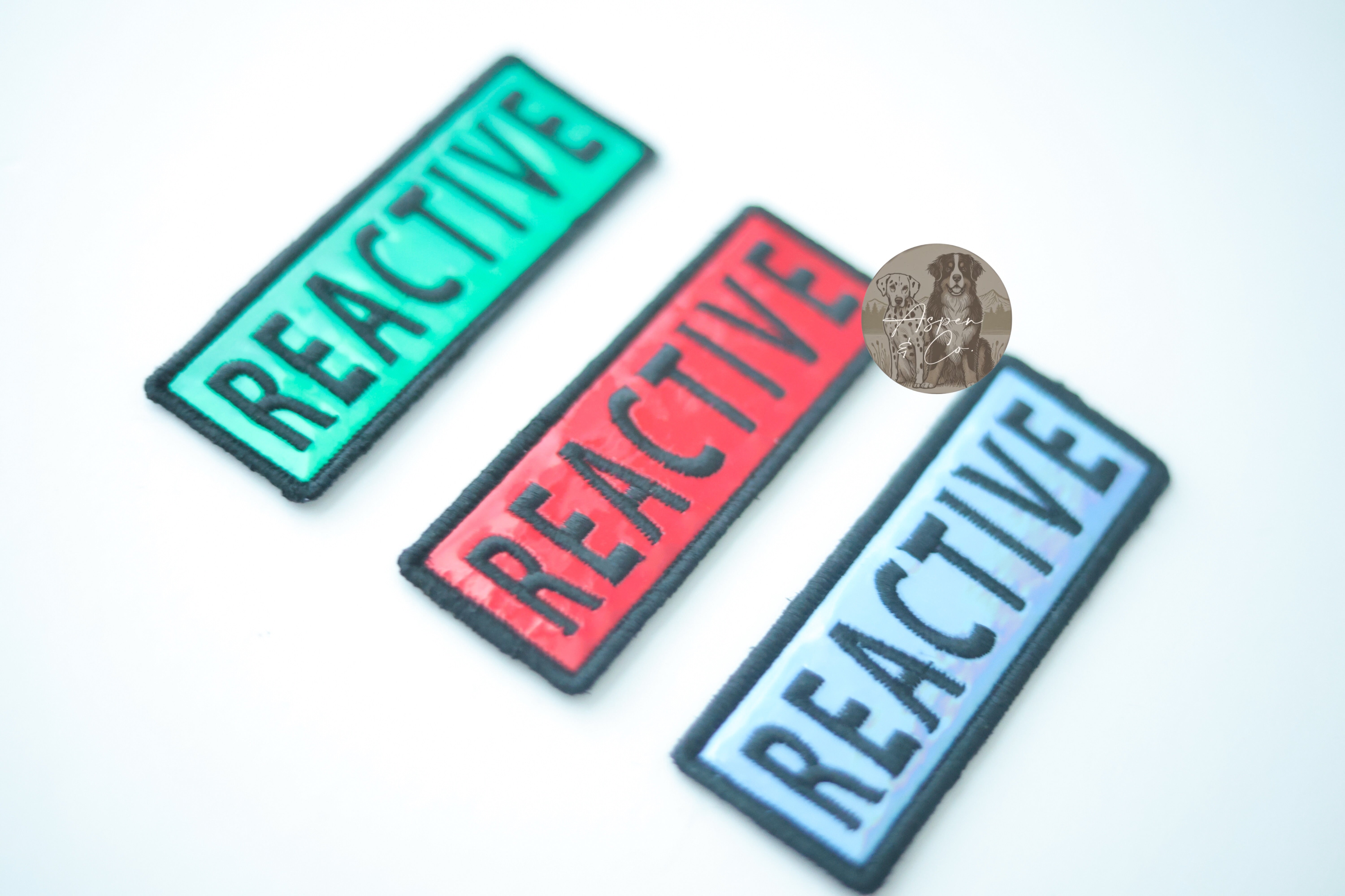 Photo displaying holographic dog patches with the word "Reactive" in multiple color options, perfect for attaching to collars, harnesses, vests, or capes.