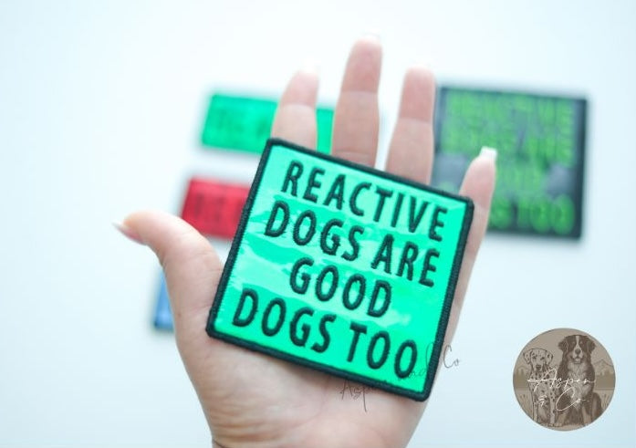 Close-up photo of a greenk holographic dog patch with neon green text reading "Reactive Dogs Are Good Dogs Too," designed for harnesses, vests, or capes.