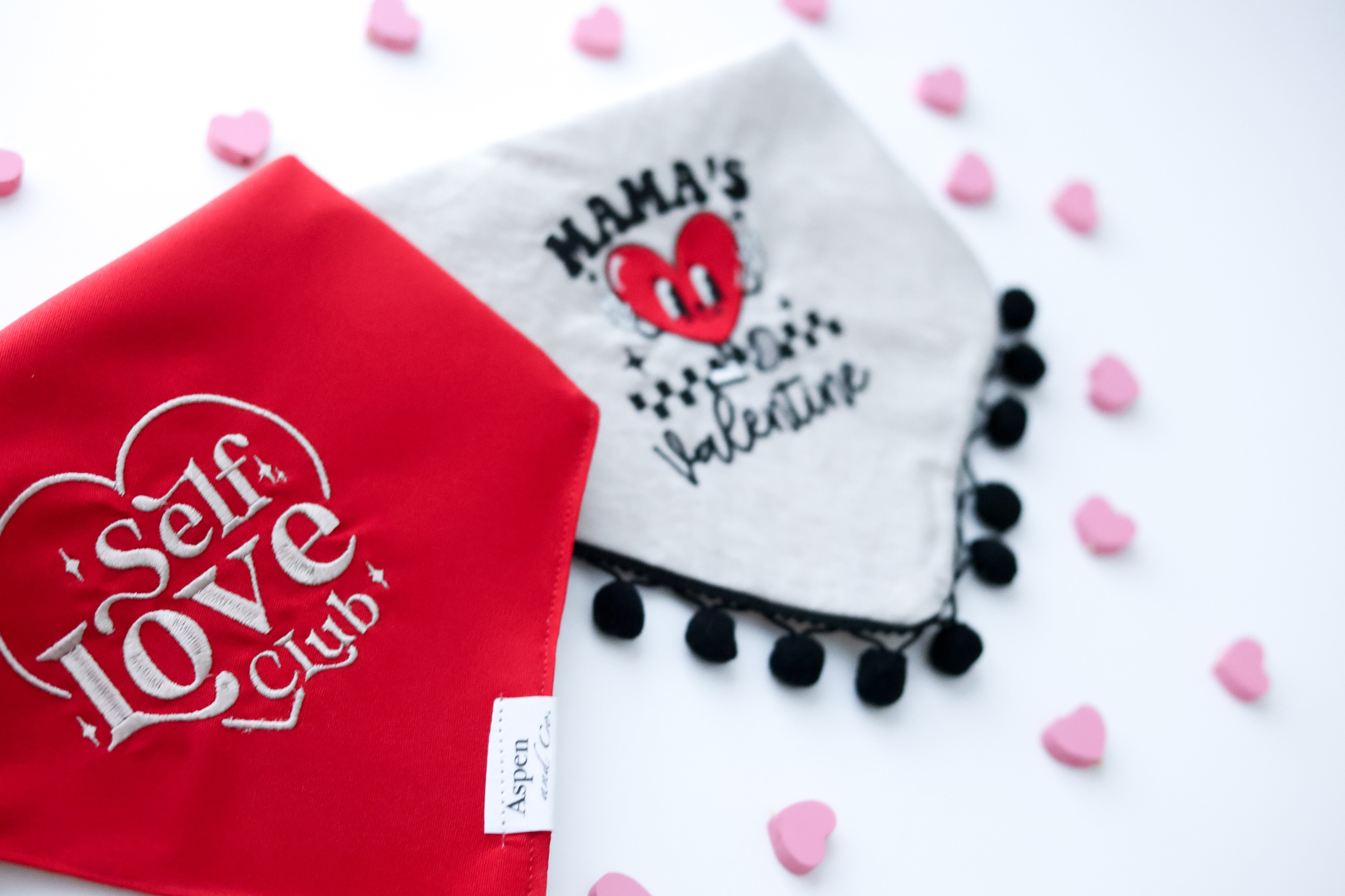 Two embroidered dog bandanas side by side—one cool-toned red bandana with beige stitching that says “Self Love Club” inside a heart, and one pink linen bandana with red embroidery reading “Mama’s Valentine”; both are stylish Valentine’s Day accessories for dogs.